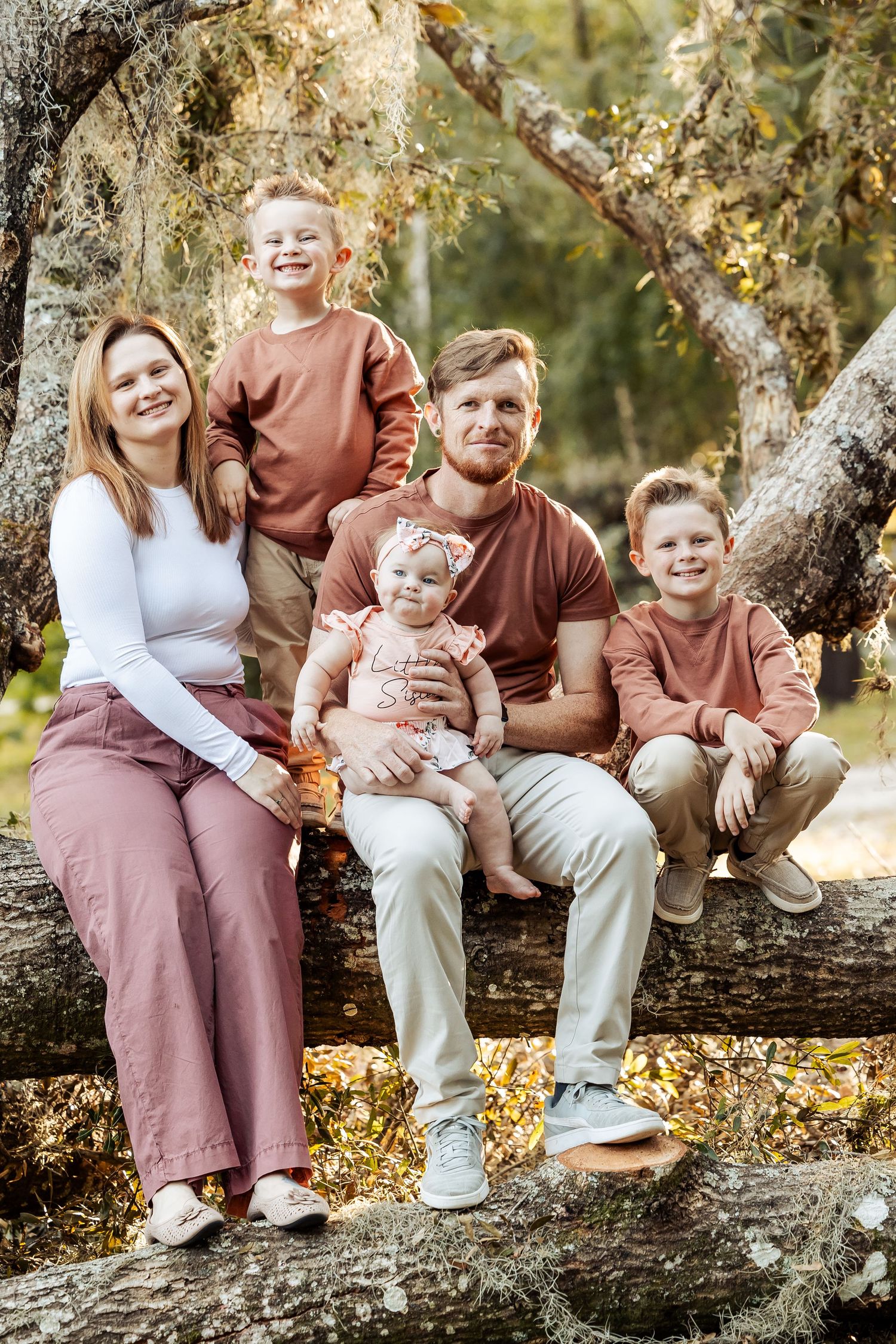 Family of five sitting on a fallen log outdoors, dressed in coordinating rust and pink tones.