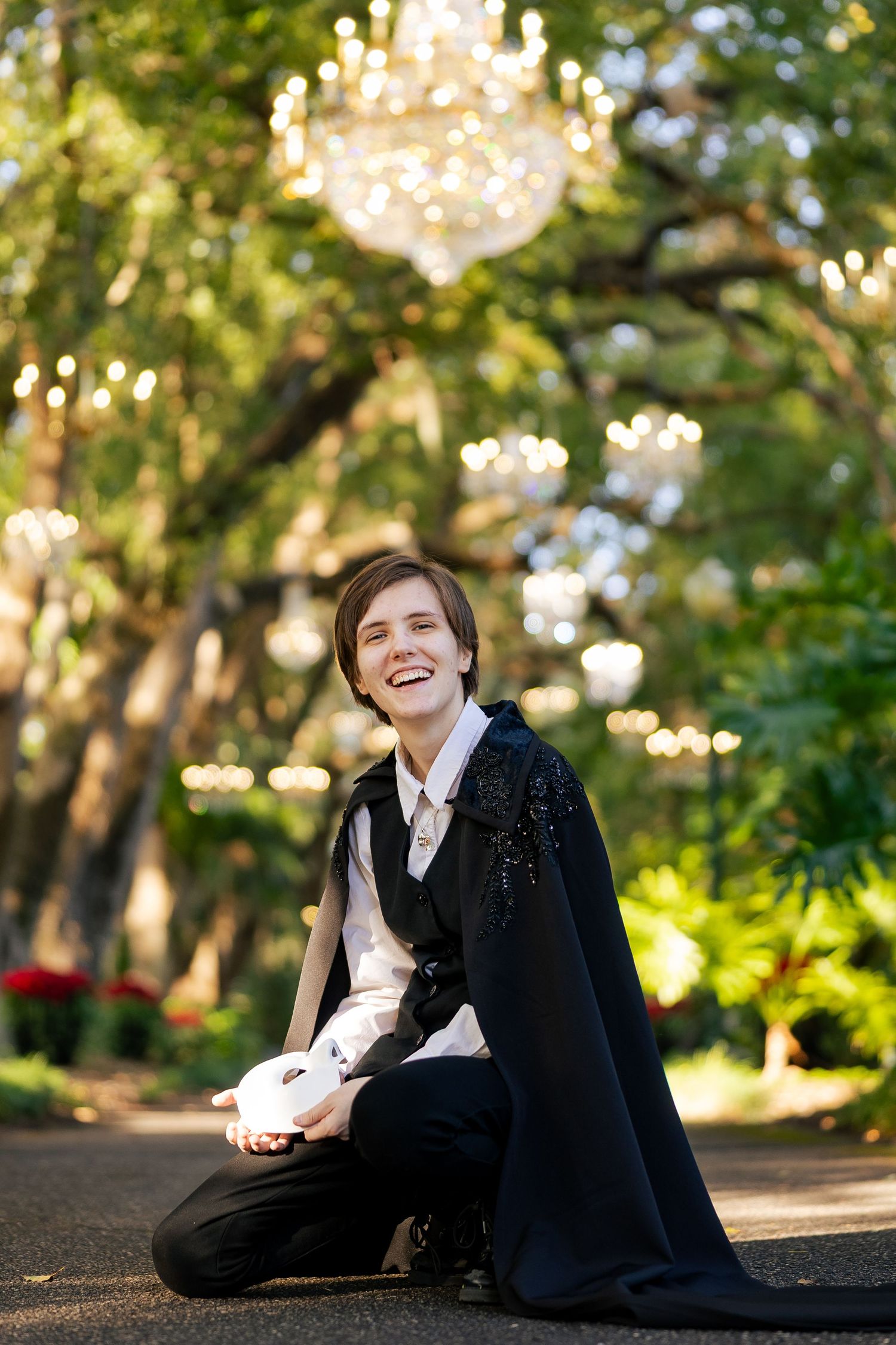 Person in black cape costume kneeling outdoors, laughing joyfully, holding white mask under chandelier lights.