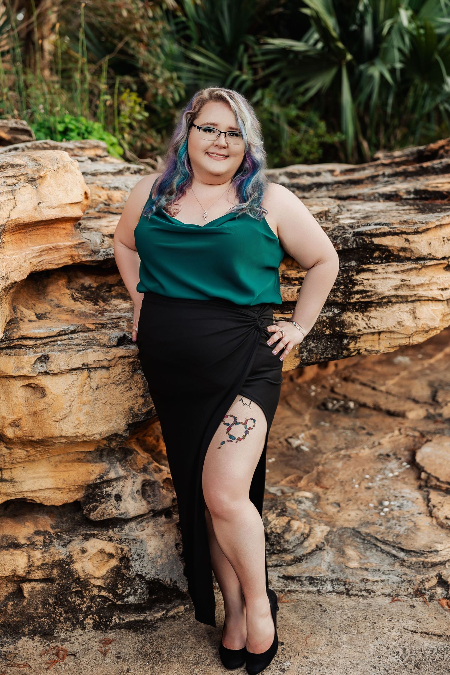 Woman with colorful hair in green top and black slit skirt poses against sandstone rocks in lush garden.