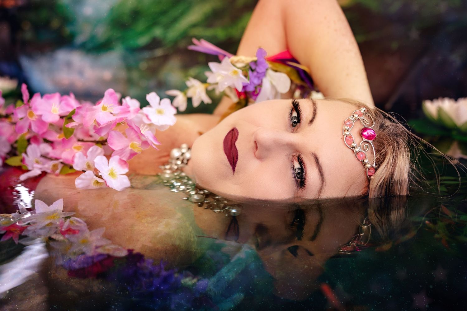Woman lying in water surrounded by pink and purple flowers wearing ornate golden headpiece.