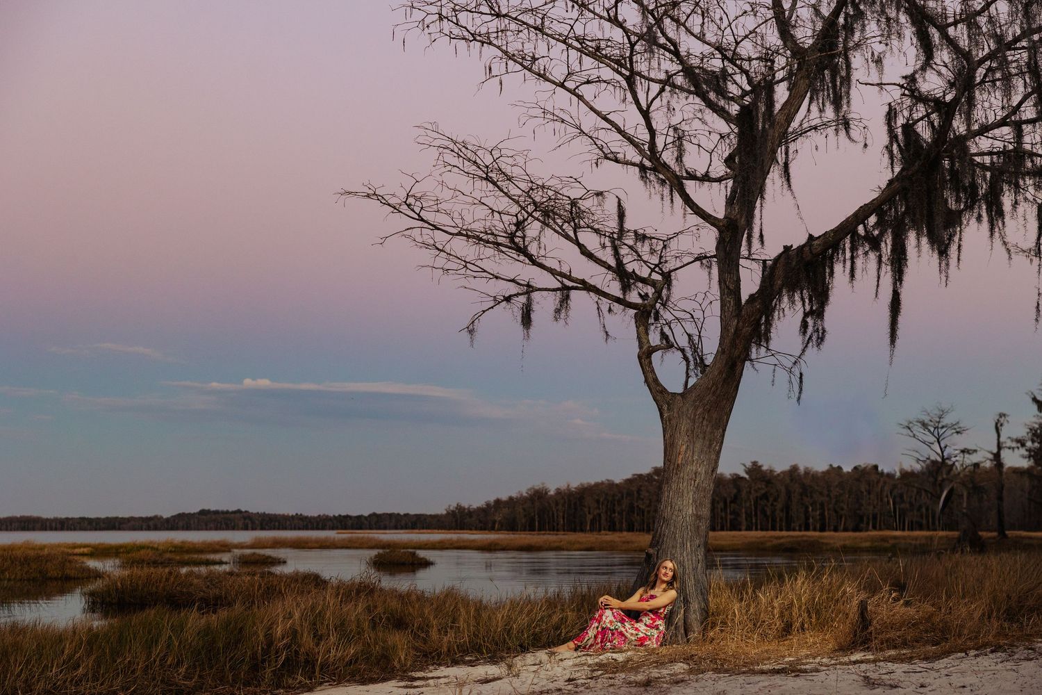 Woman in red dress resting under a moss-draped tree at sunset by a serene coastal marsh.