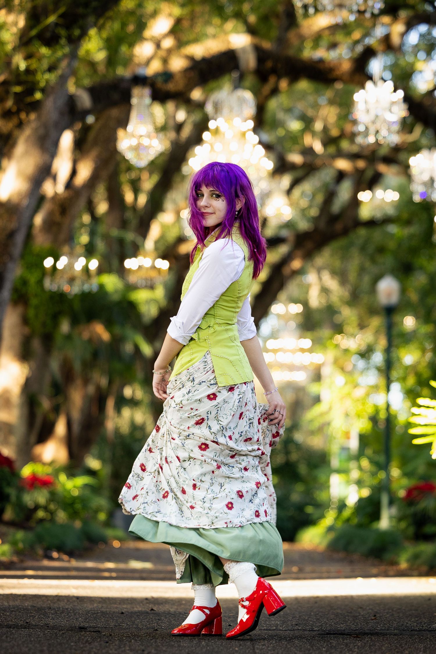 Woman in purple hair, green vest, floral skirt, and red heels poses in a chandelier-lit garden path.