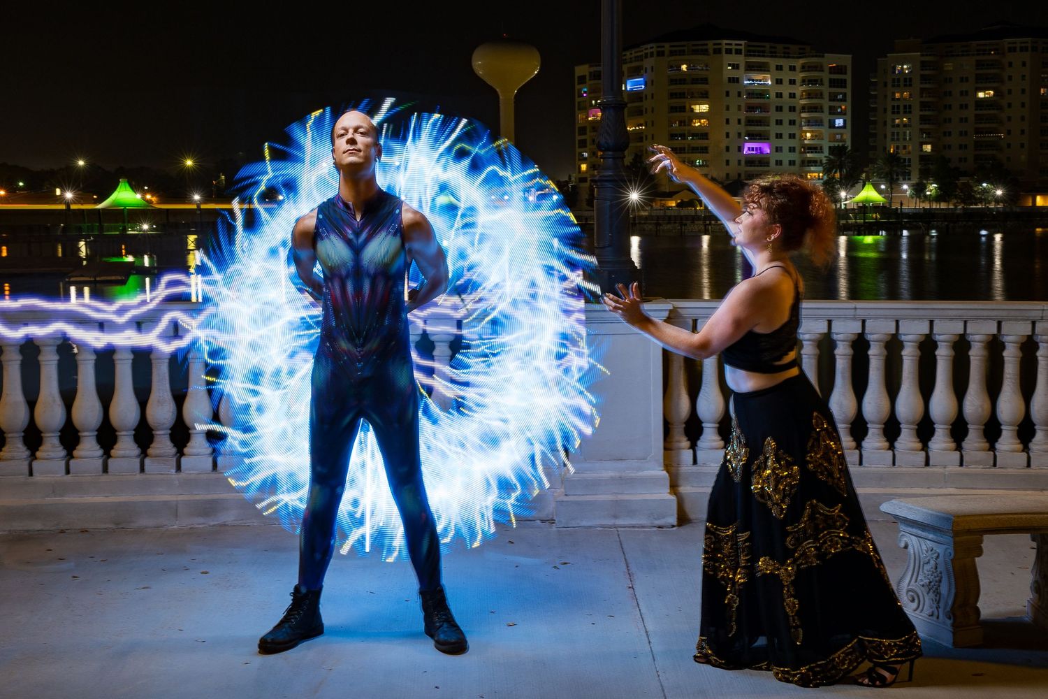 Two cosplay performers create a dramatic light painting scene with electric blue energy effects at night by a waterfront.