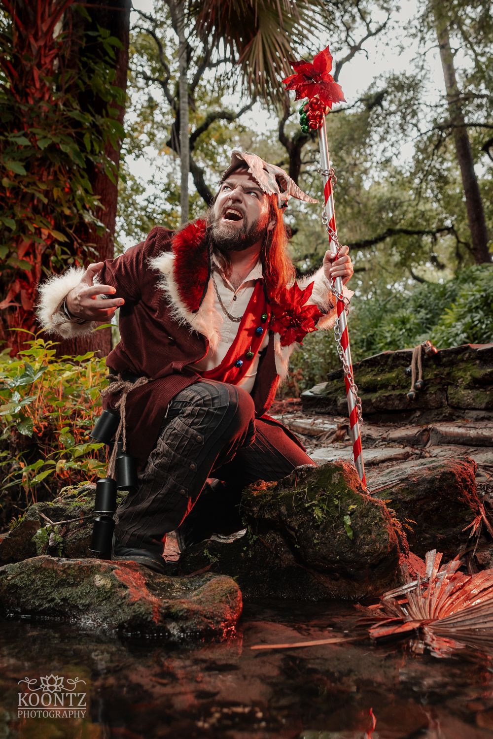 A fantasy cosplayer in a red fur-trimmed coat crouches on rocks holding a red floral staff in an outdoor forest setting.