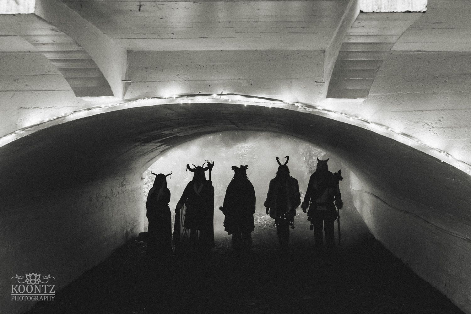Black and white silhouette of five costumed figures with horns standing in a tunnel archway.