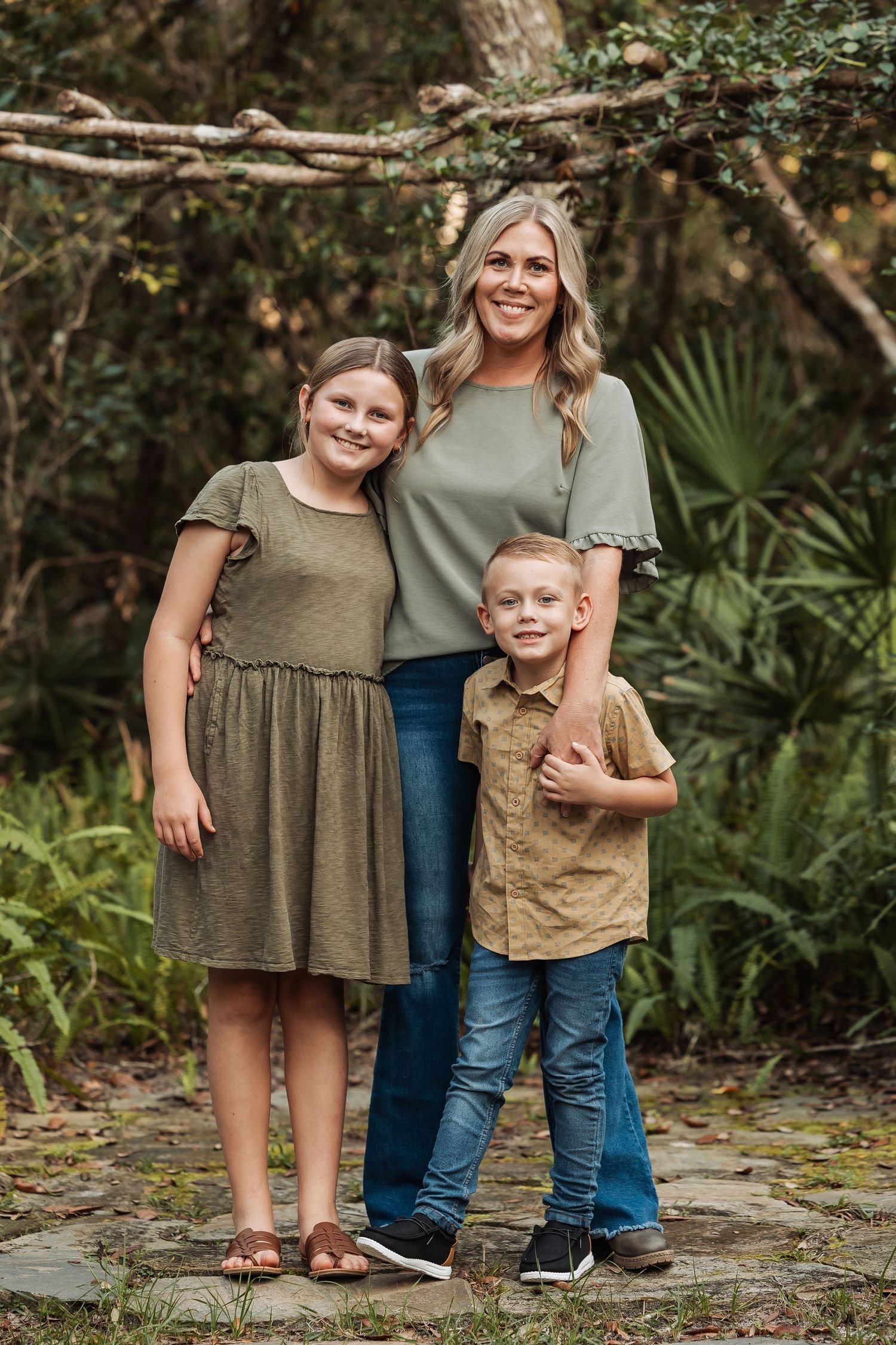 A blonde mother smiles with her two children in a lush green outdoor nature setting.