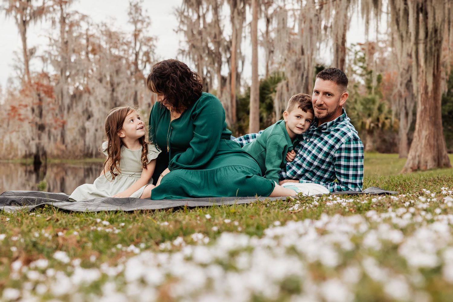 Family of four sitting together in a flower field near a lake with cypress trees.