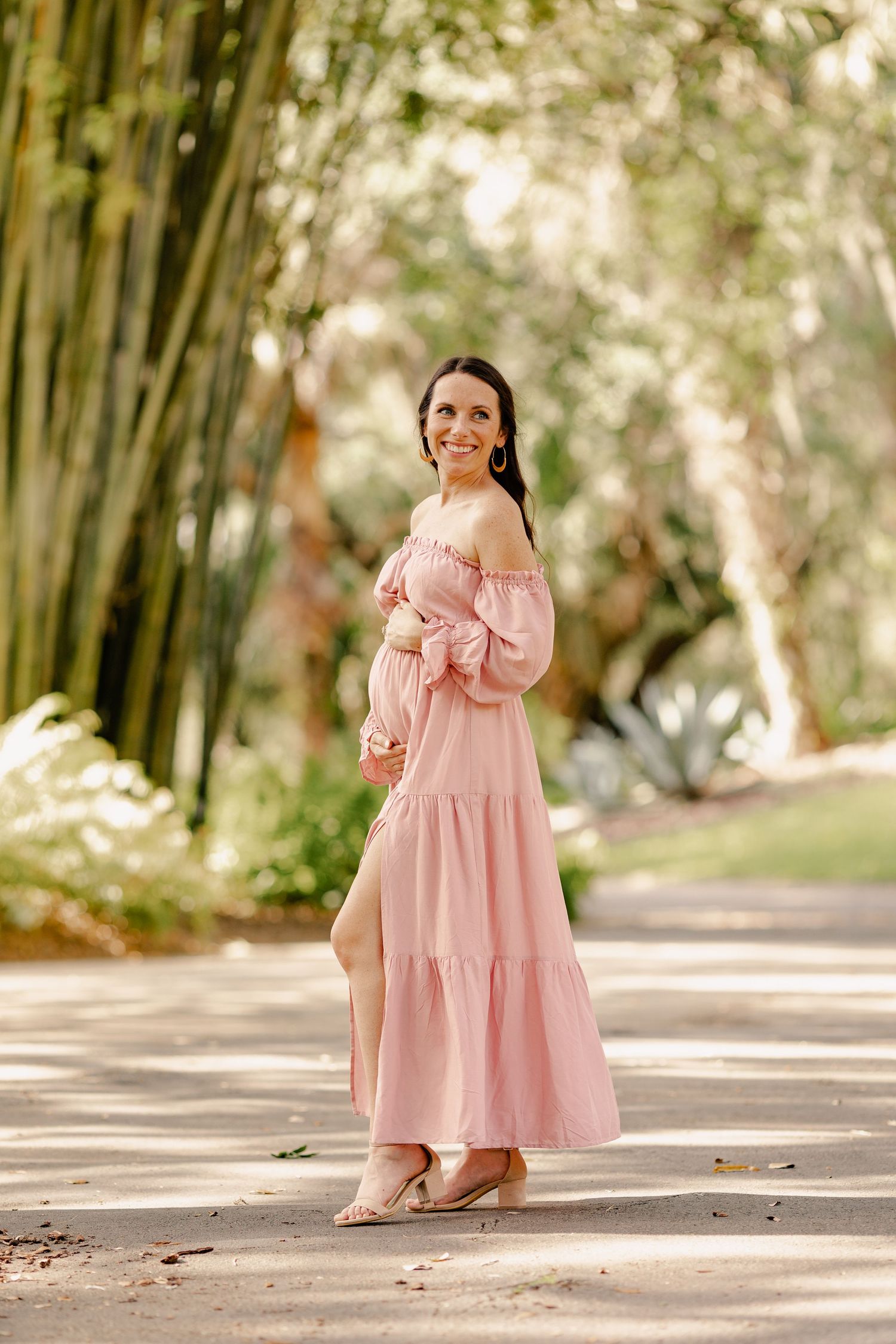 Smiling pregnant woman in pink off-shoulder maxi dress walking through bamboo garden path.