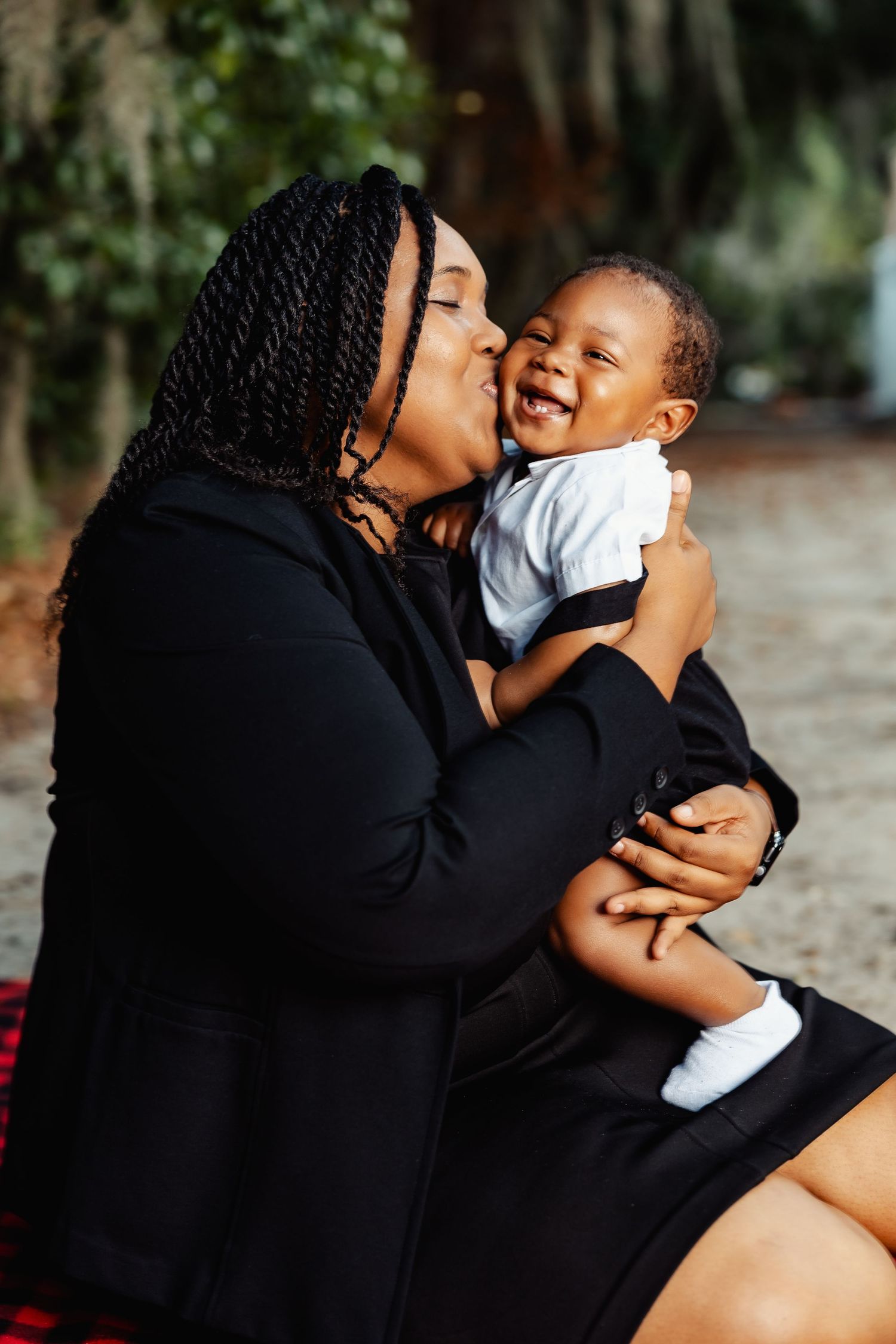 Joyful Black mother kissing laughing baby boy while sitting outdoors under shaded trees.