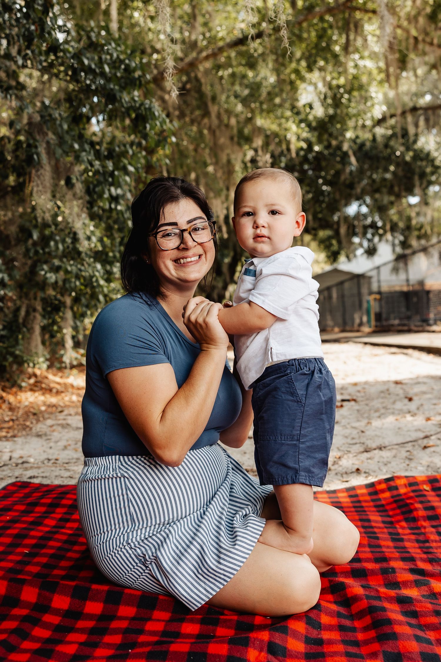 A smiling mother sits on a red plaid blanket holding her baby boy outdoors.