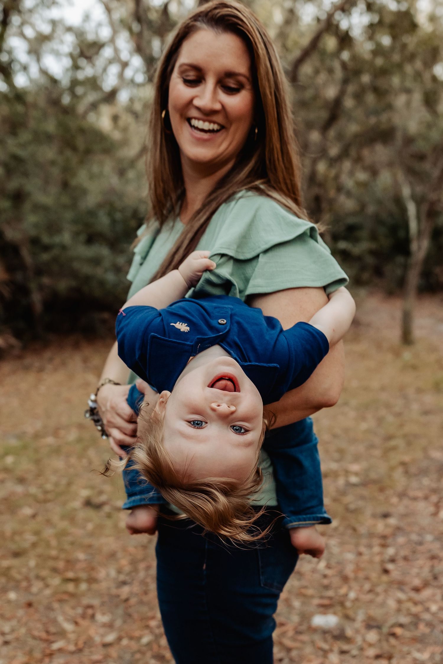Laughing mother holding toddler upside down playfully in a wooded outdoor setting.