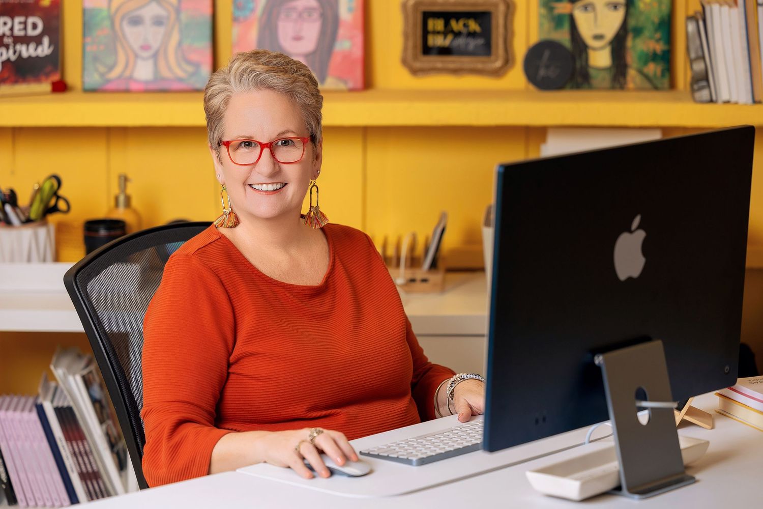 Smiling woman with red glasses and orange top works at an Apple iMac desktop computer in a colorful office.