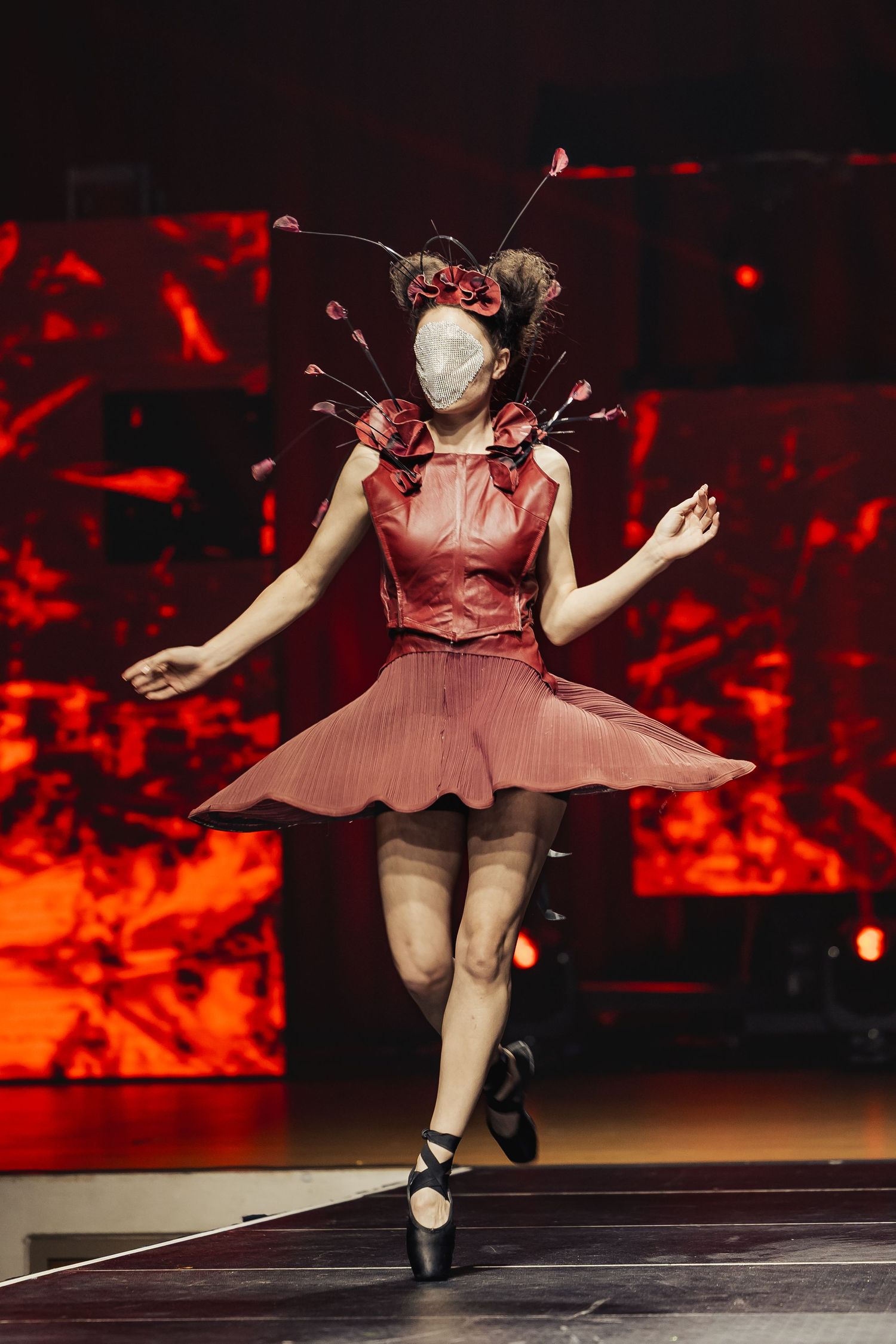Dancer in red floral costume with face mask and hair accessories performs ballet on stage with red lighting.