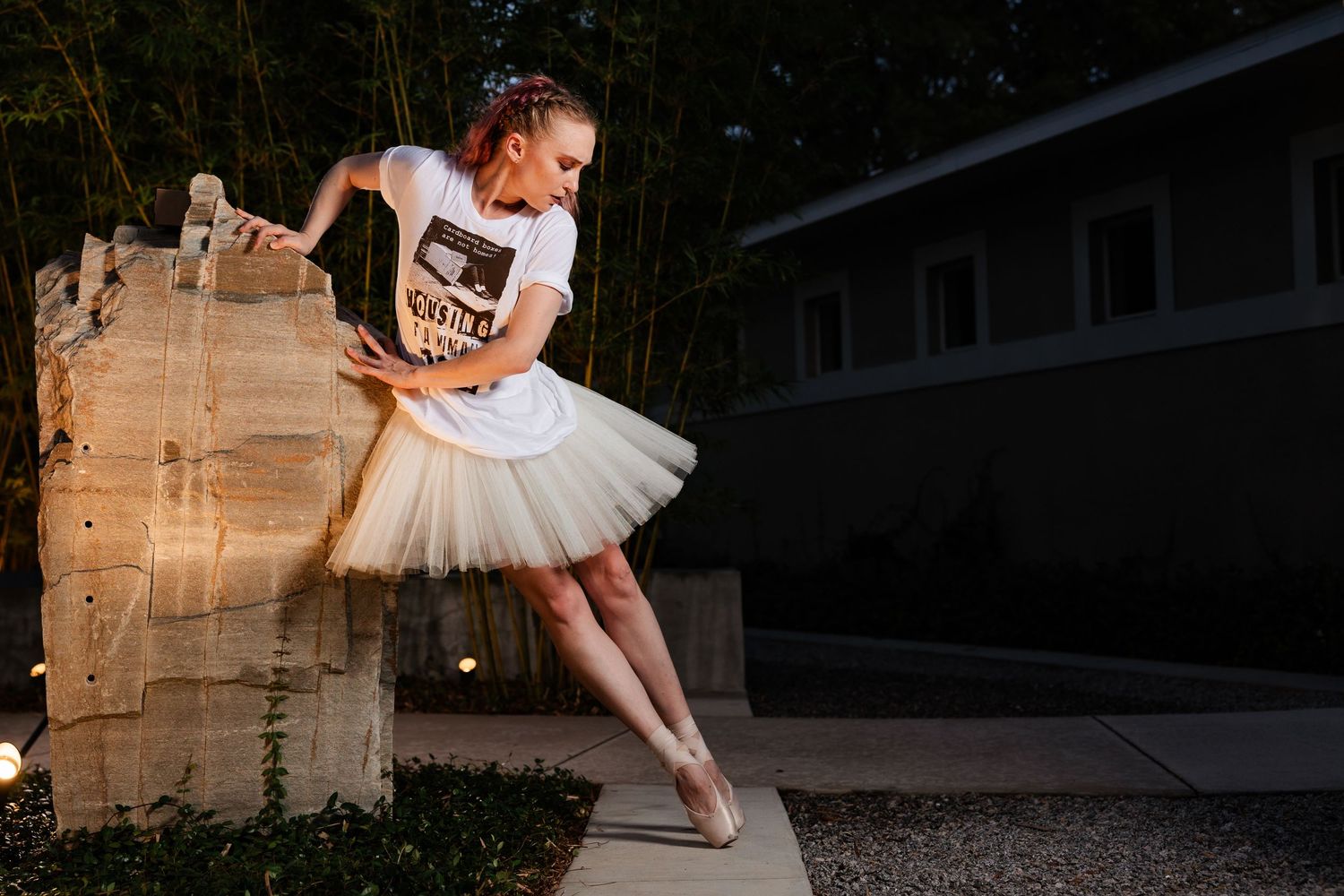 Ballerina in white tutu and graphic tee performing an elegant pose on a stone structure outdoors.