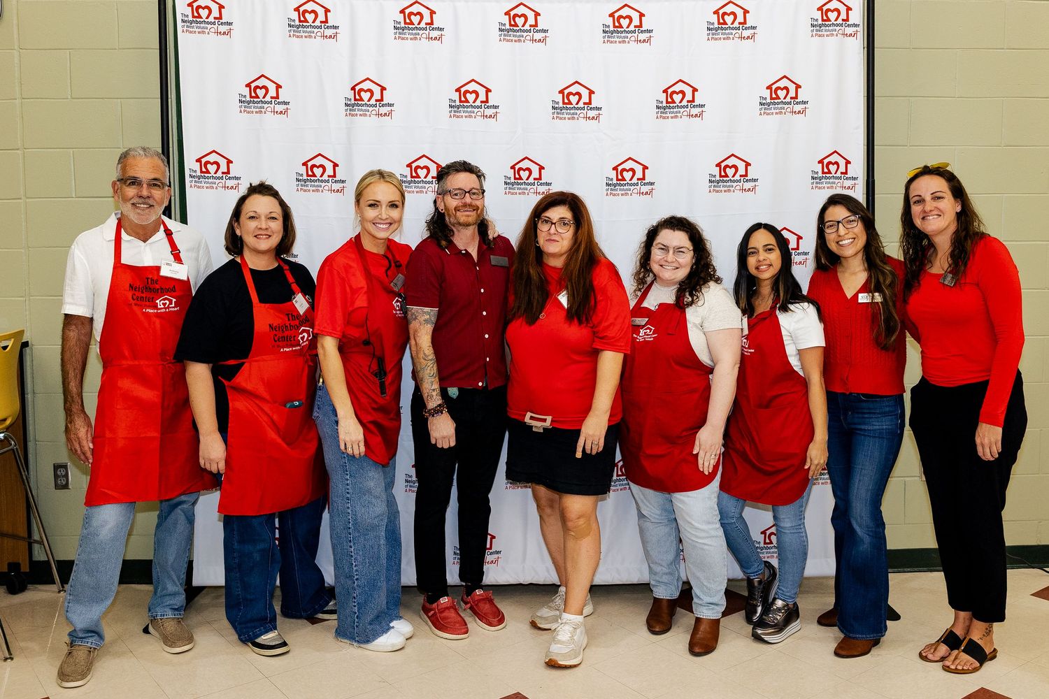 Team of volunteers in red aprons smiling together in front of a charity organization branded backdrop.