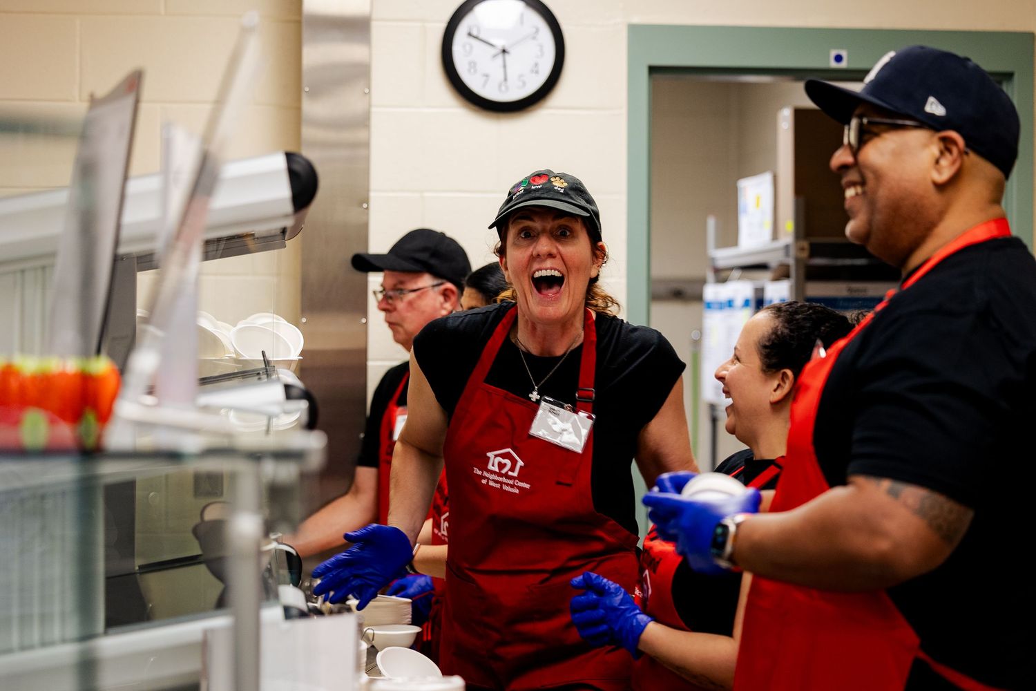 Enthusiastic kitchen volunteers in red aprons laugh together while preparing food at a community event.