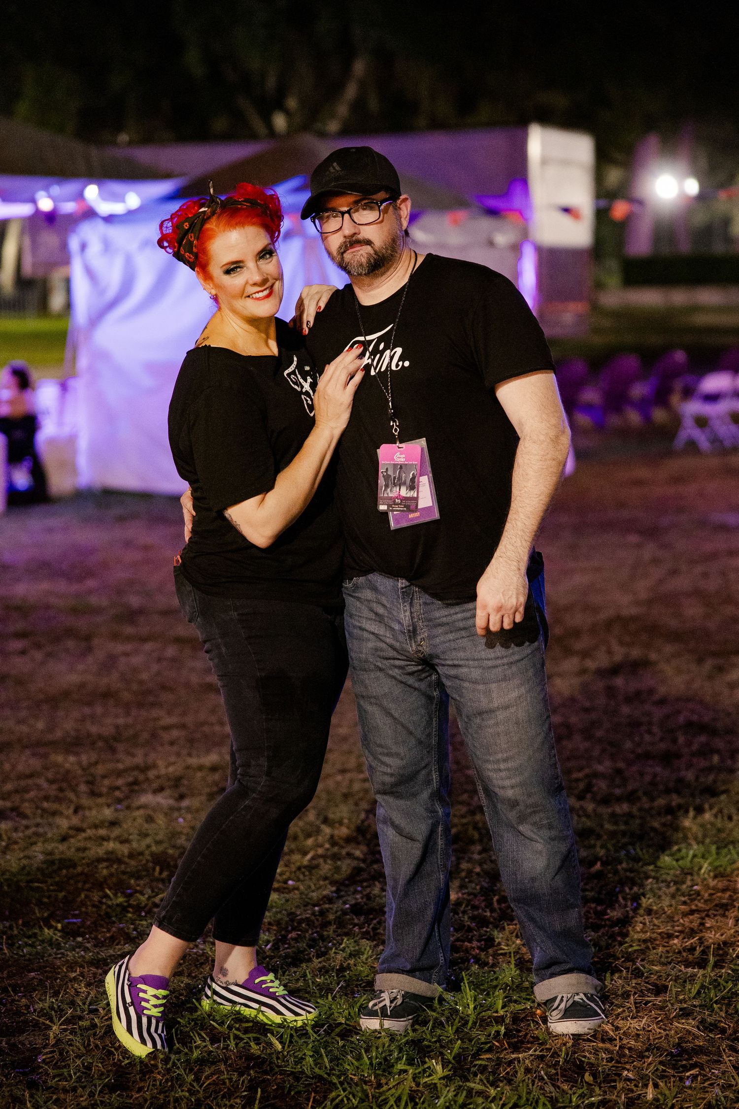 Couple posing together at an outdoor night festival, woman with red hair in black outfit.