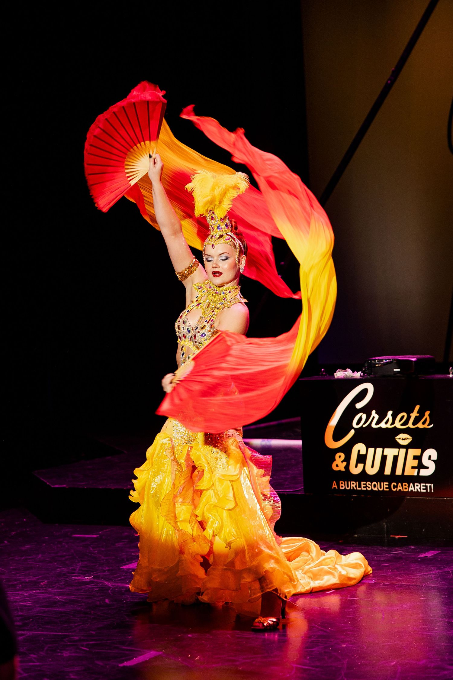 Burlesque dancer waves red and yellow silk fans in ornate golden costume at Corsets and Cuties cabaret.