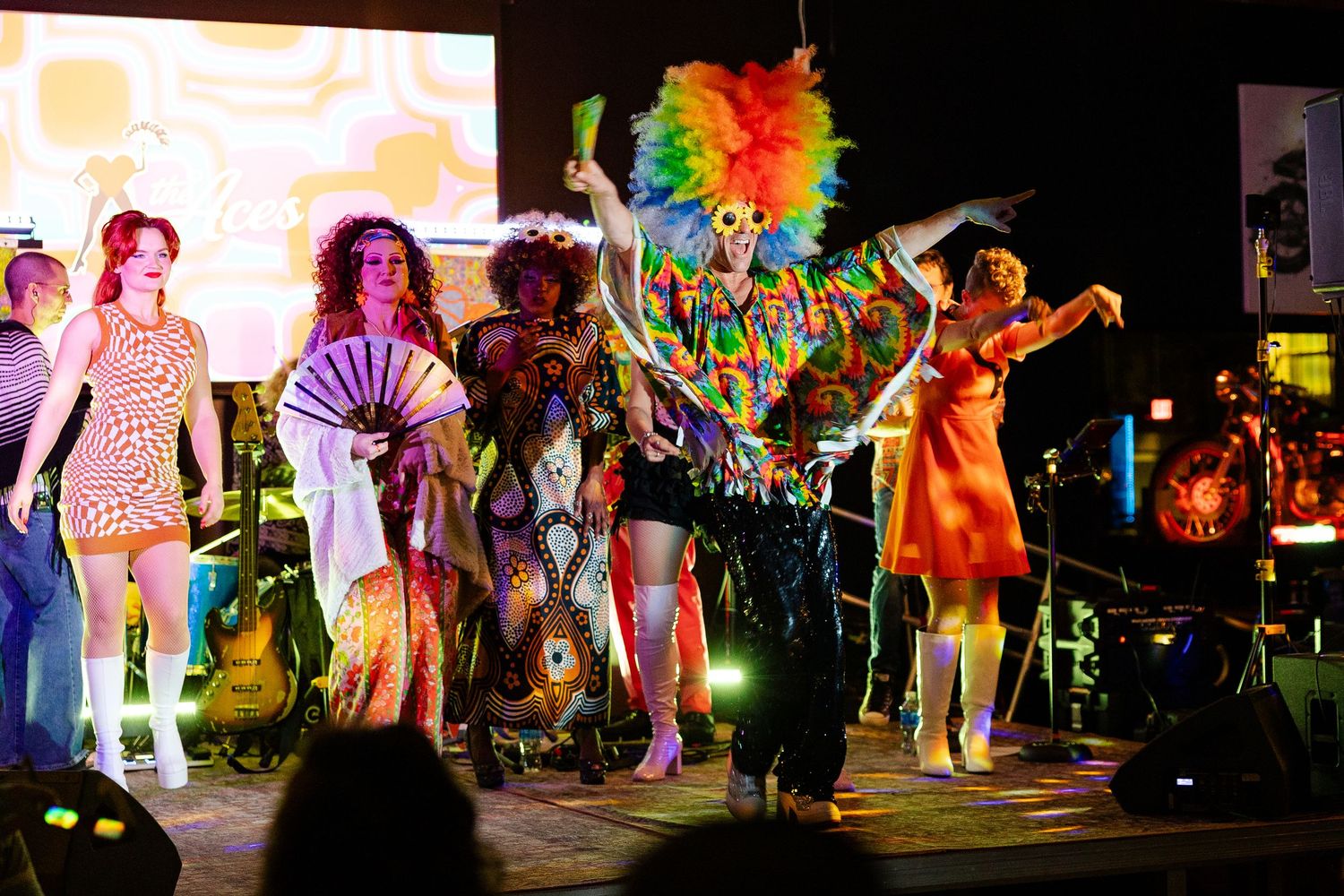 Colorful drag performers dancing on an outdoor stage with a band performing behind them at night.