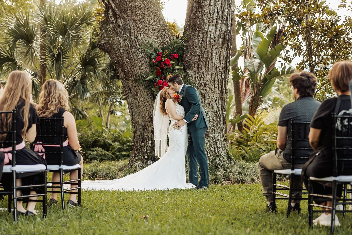 Bride and groom share first kiss at outdoor wedding ceremony beneath large tree with red floral wreath.