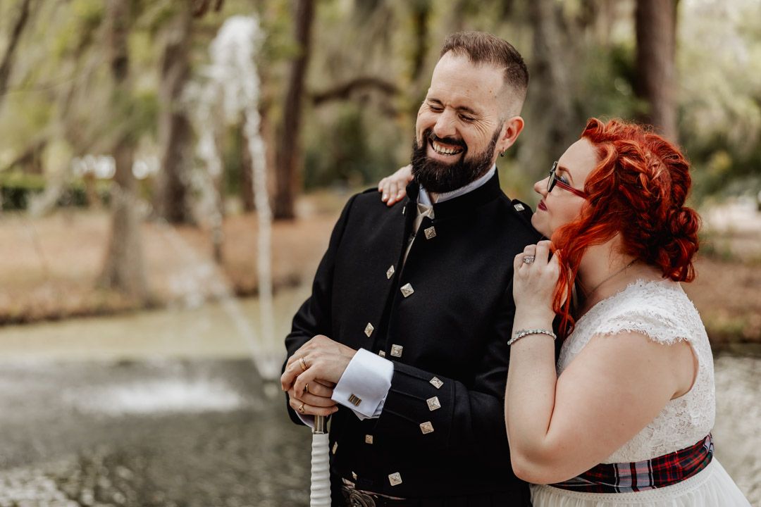 Smiling groom in Scottish military jacket and bride in white dress laughing together near a garden fountain.