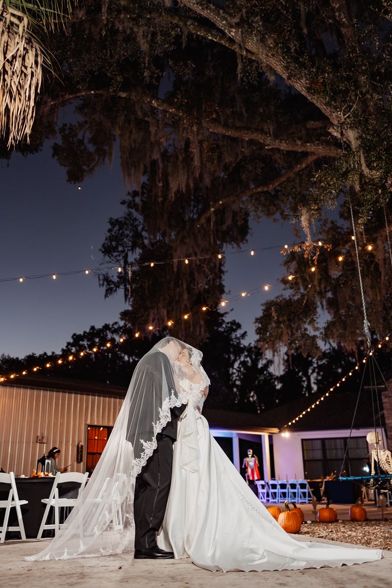Bride and groom share romantic kiss under Spanish moss tree with string lights at outdoor night wedding.