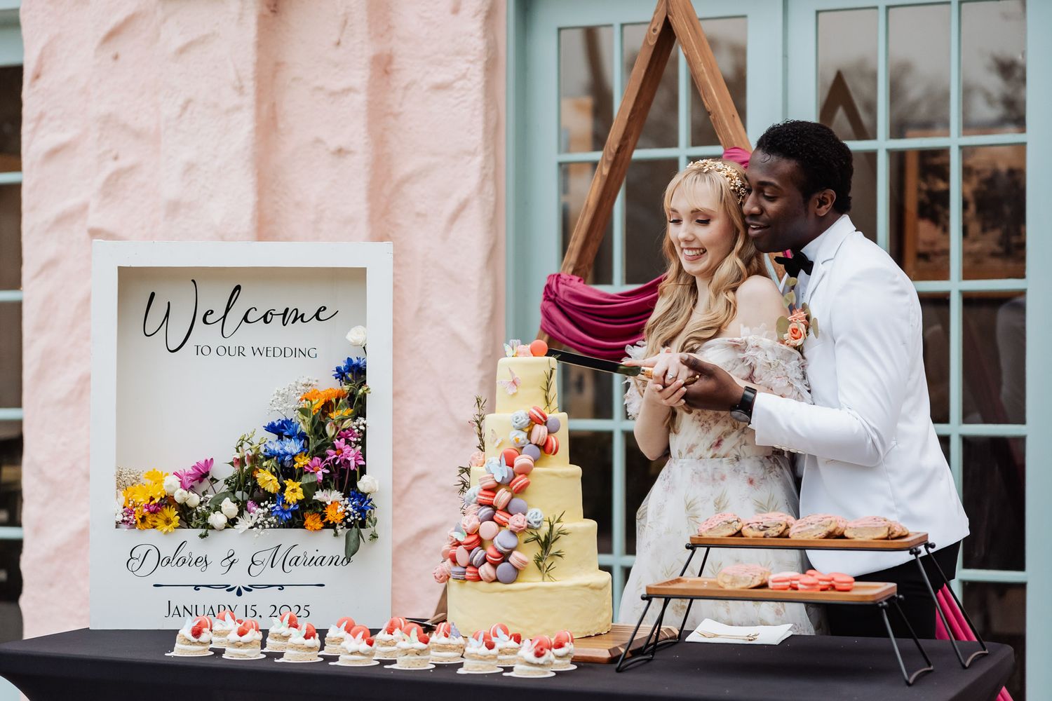 Interracial couple cutting colorful wedding cake beside welcome sign and dessert table display.