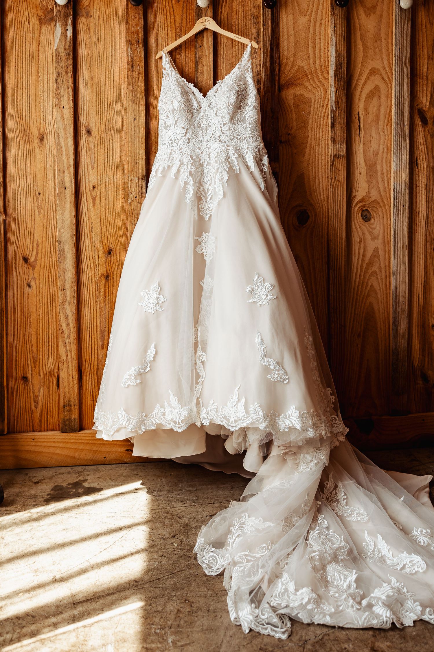 Elegant white lace wedding gown with long train hanging against wooden paneled wall.