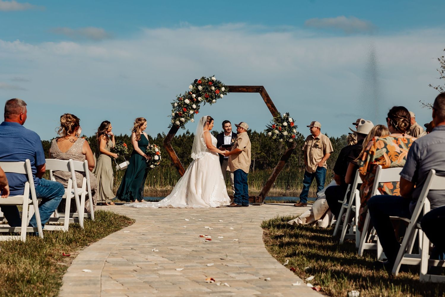 Outdoor wedding ceremony under a floral arch with guests seated on white chairs along a stone pathway.