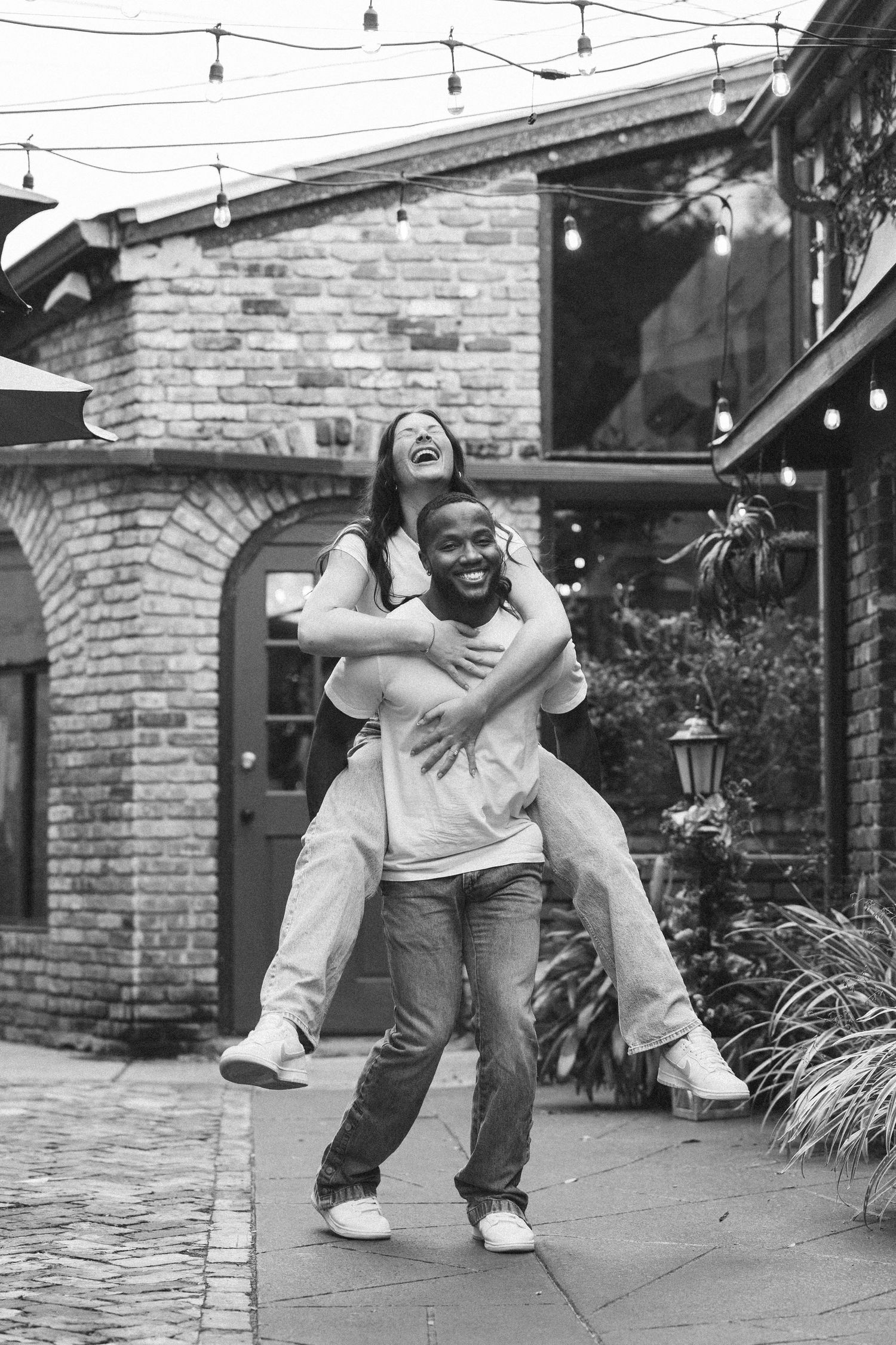 Black and white shot of couple having fun with piggyback ride in brick courtyard.