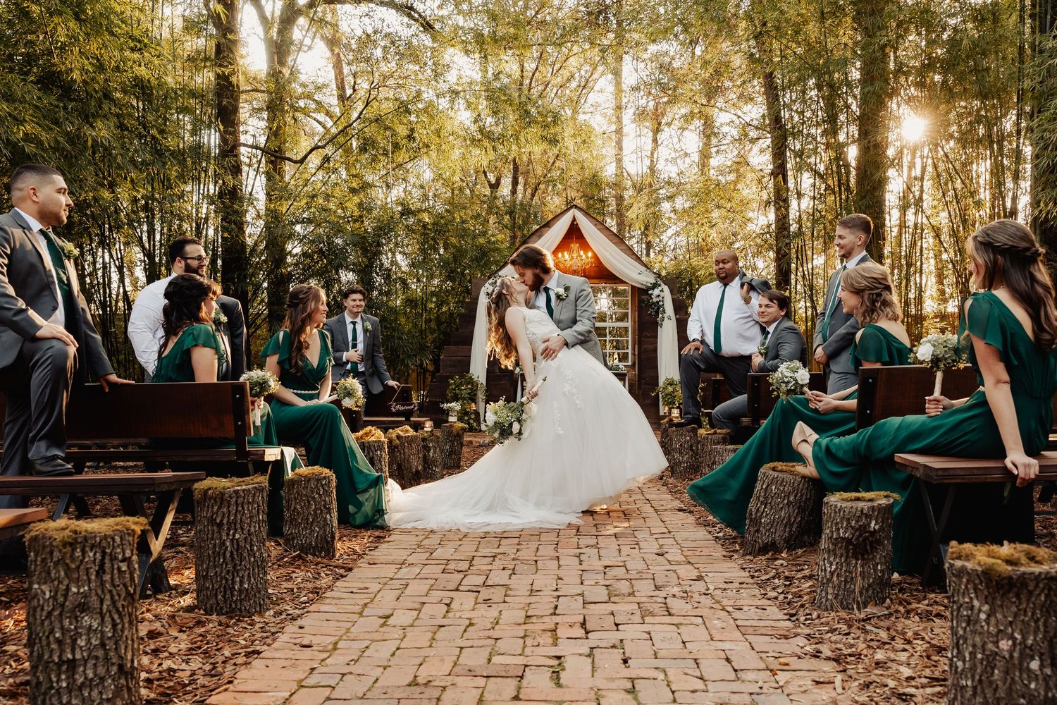 Wedding ceremony seating arrangement on brick pathway with wooden stump benches in forest.