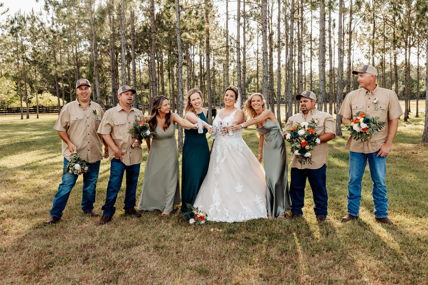 Wedding party poses outdoors with bridesmaids in elegant dresses and groomsmen in casual tan shirts.