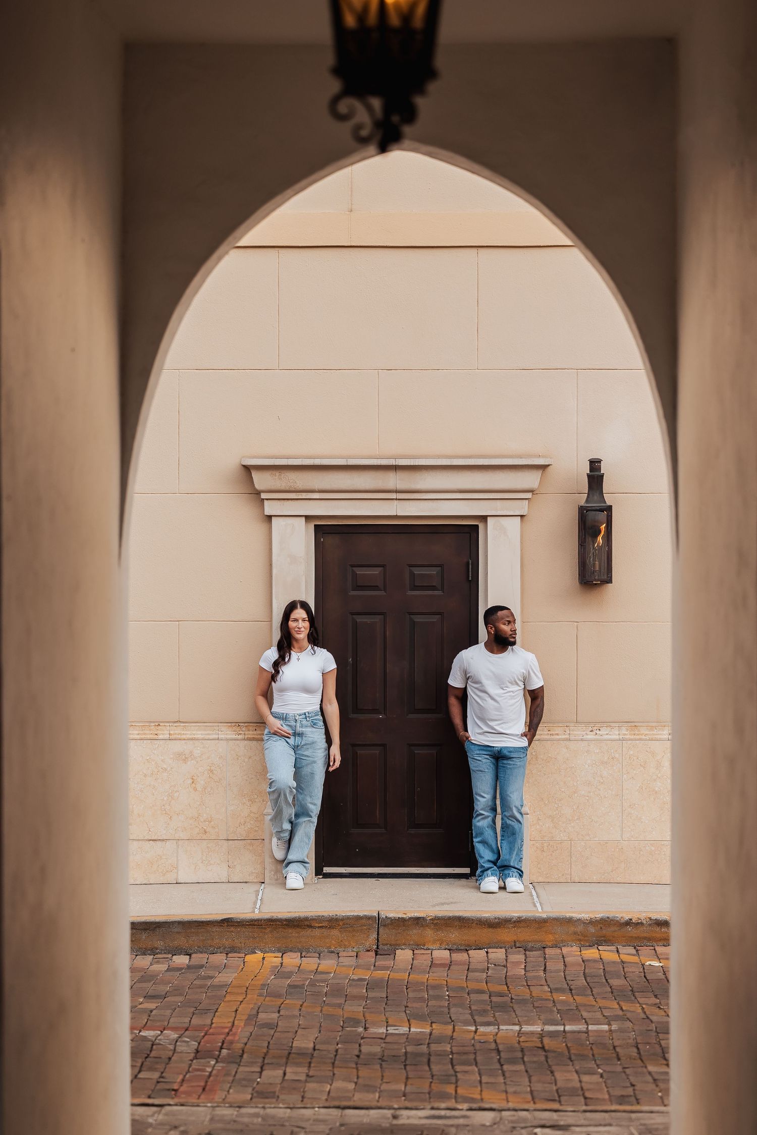 A couple in matching white shirts and jeans poses separately against a cream wall and brown door.