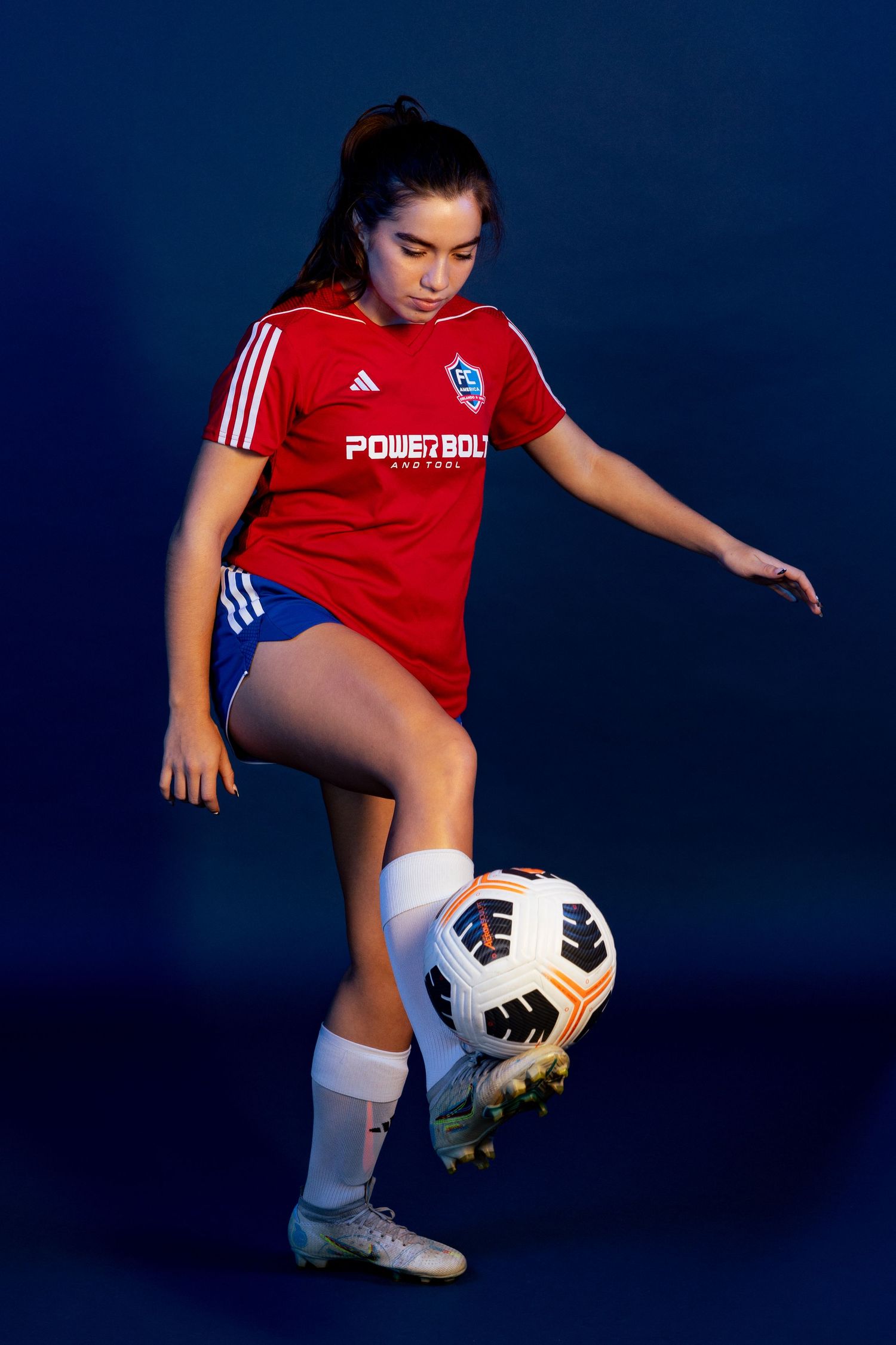 Soccer player in red Adidas jersey and blue shorts practicing ball control skills against dark blue background.