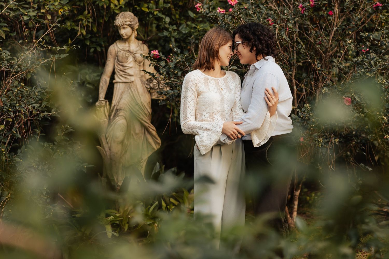Two women lean in for a kiss in a lush garden beside an elegant stone statue among pink flowers.