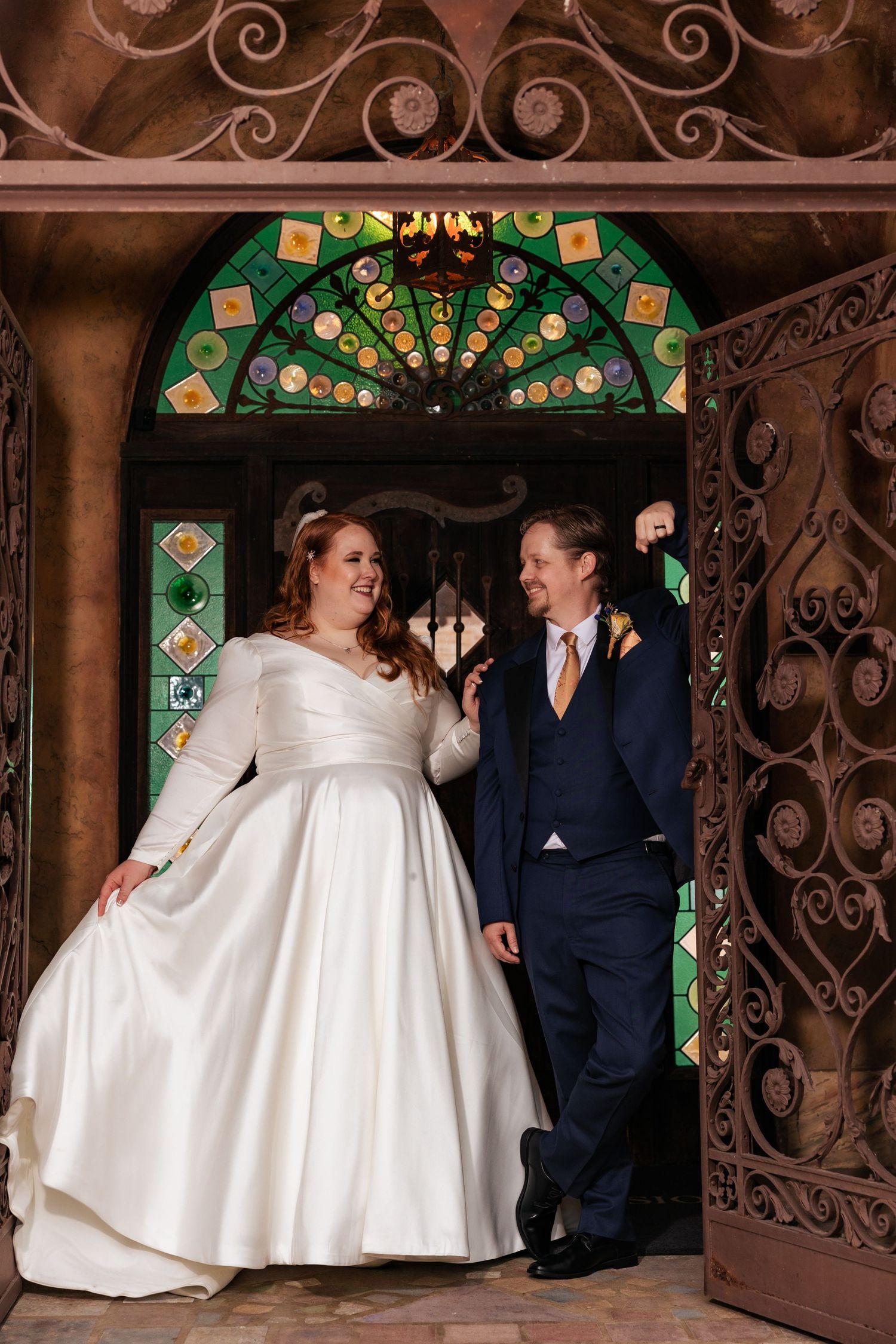 Laughing bride and groom stand in ornate iron doorway with colorful stained glass above.