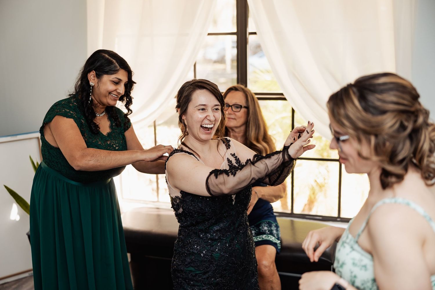 Bridesmaids laughing and helping bride get ready in elegant black lace gown before wedding ceremony.