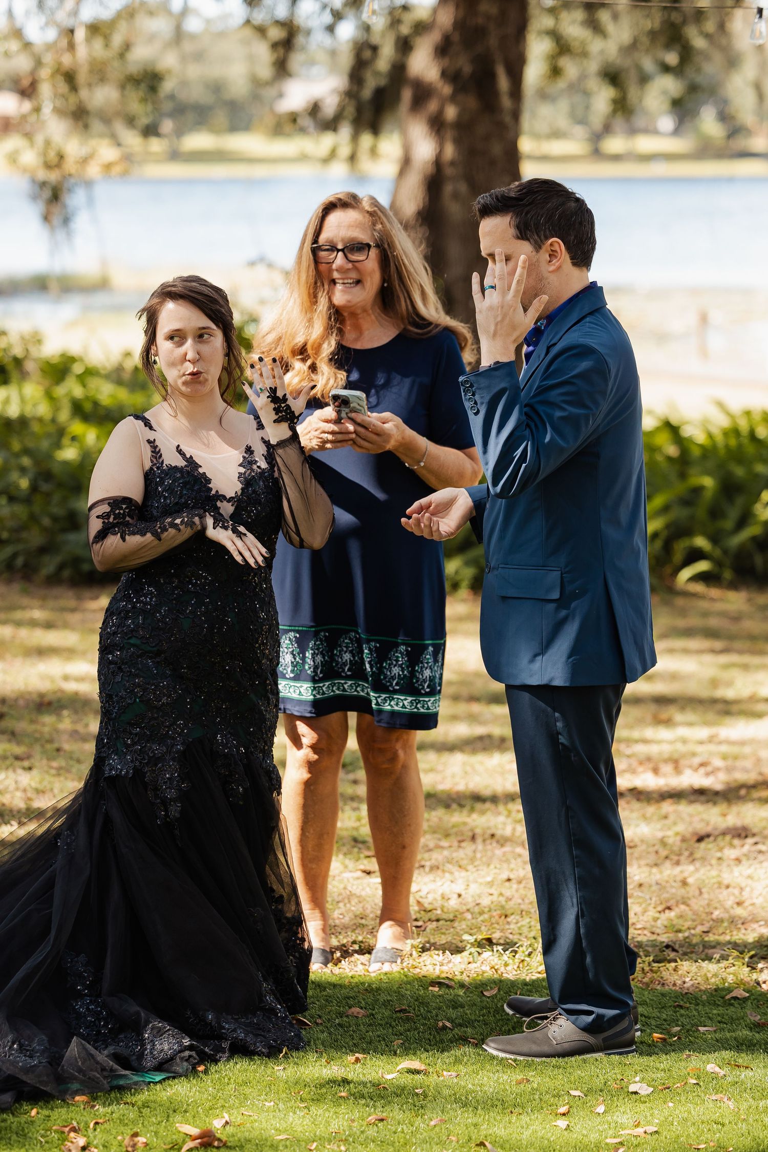 Emotional groom wipes tears during outdoor wedding ceremony as bride and officiant stand nearby under oak trees.