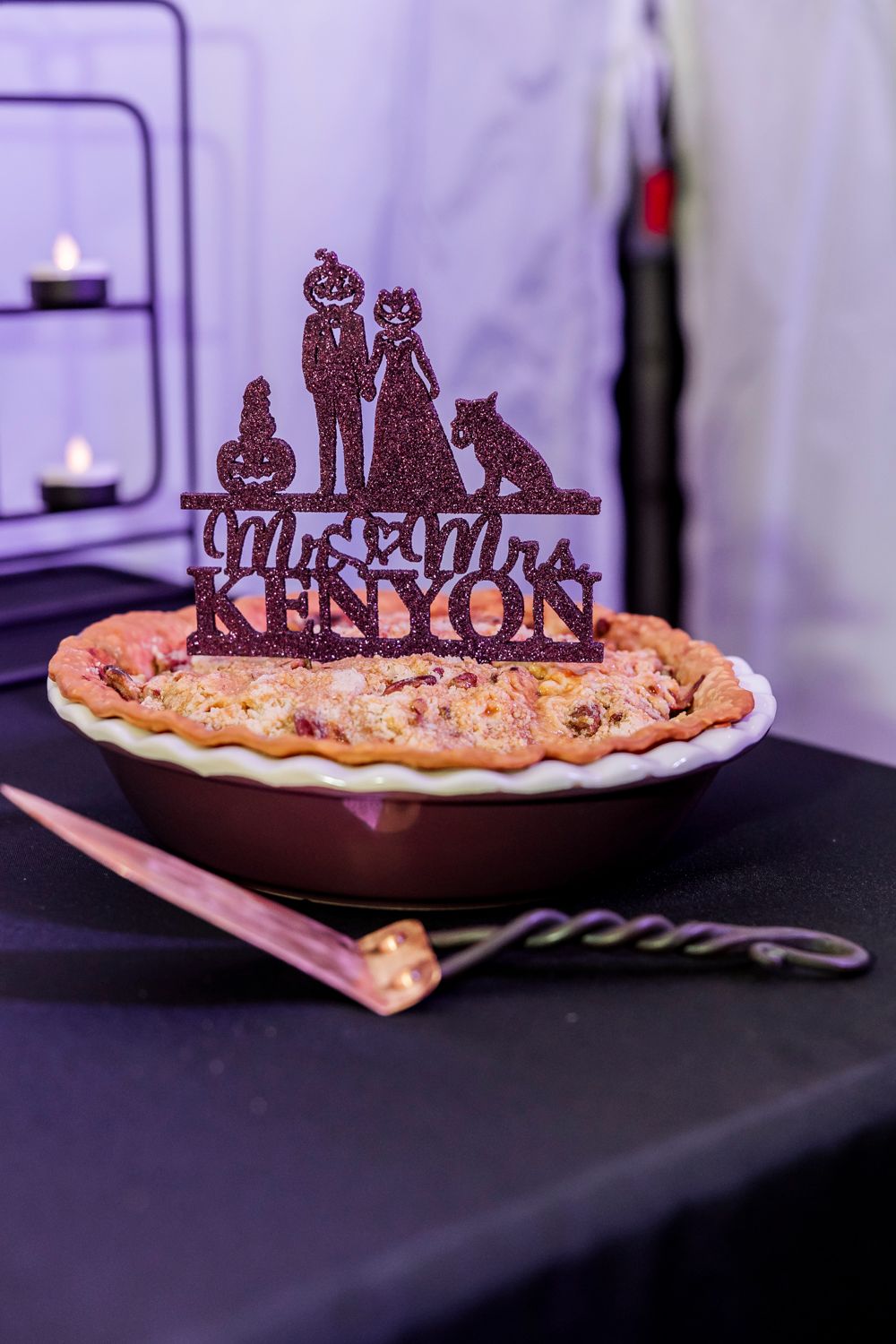 Decorative cake topper silhouette reading 'Mr & Mrs Kenyon' sits atop a rustic pie against a purple background.