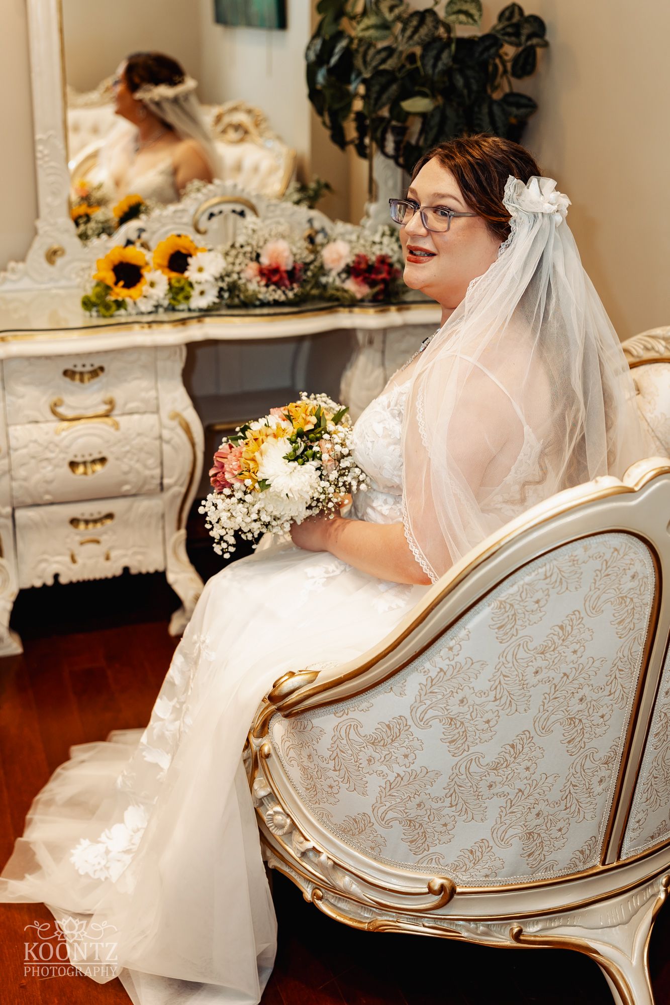 A bride sits in an ornate chair holding a sunflower bouquet next to a vintage vanity.