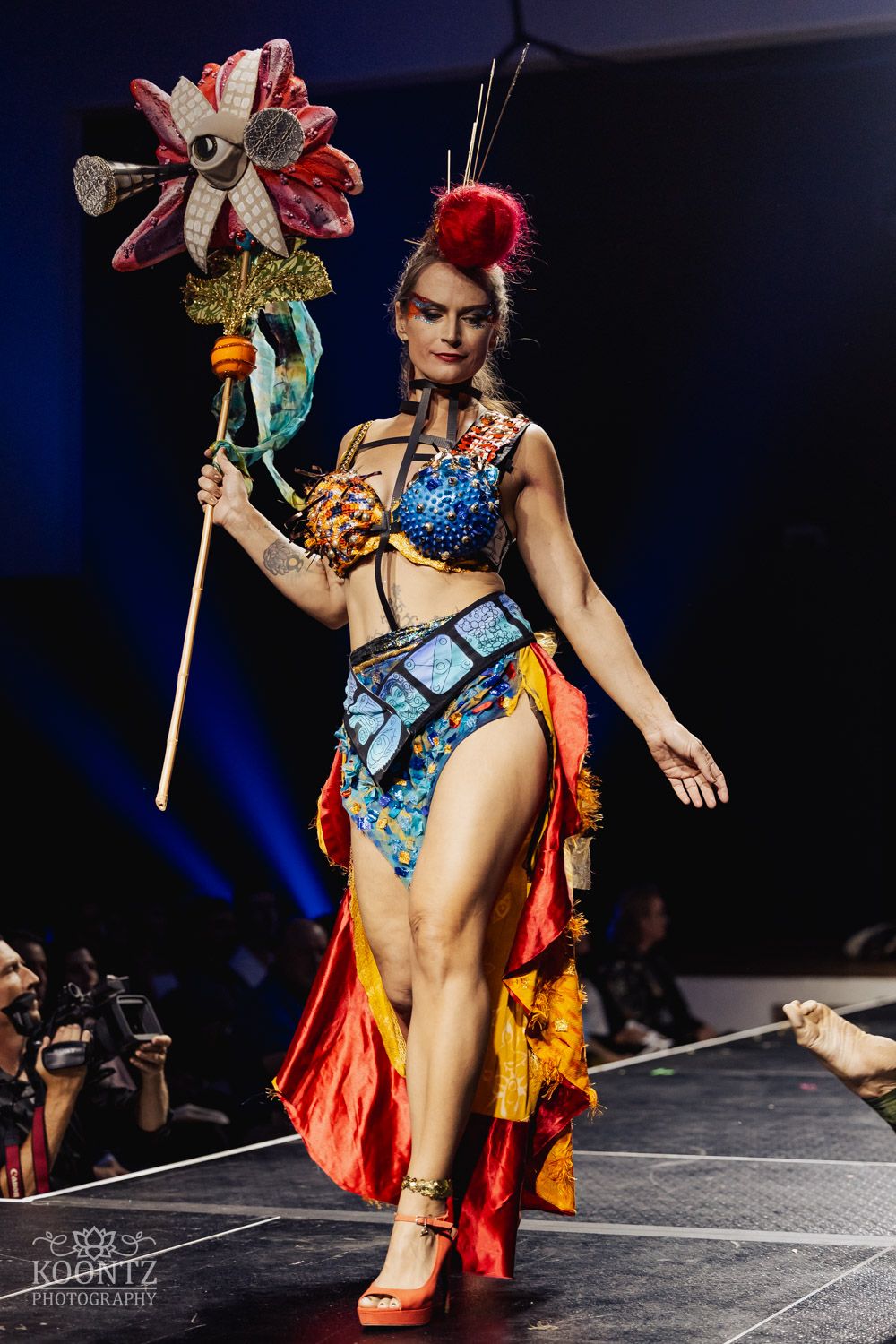 Burlesque performer in red and blue costume with feather decorations performing on dark stage during cabaret show.