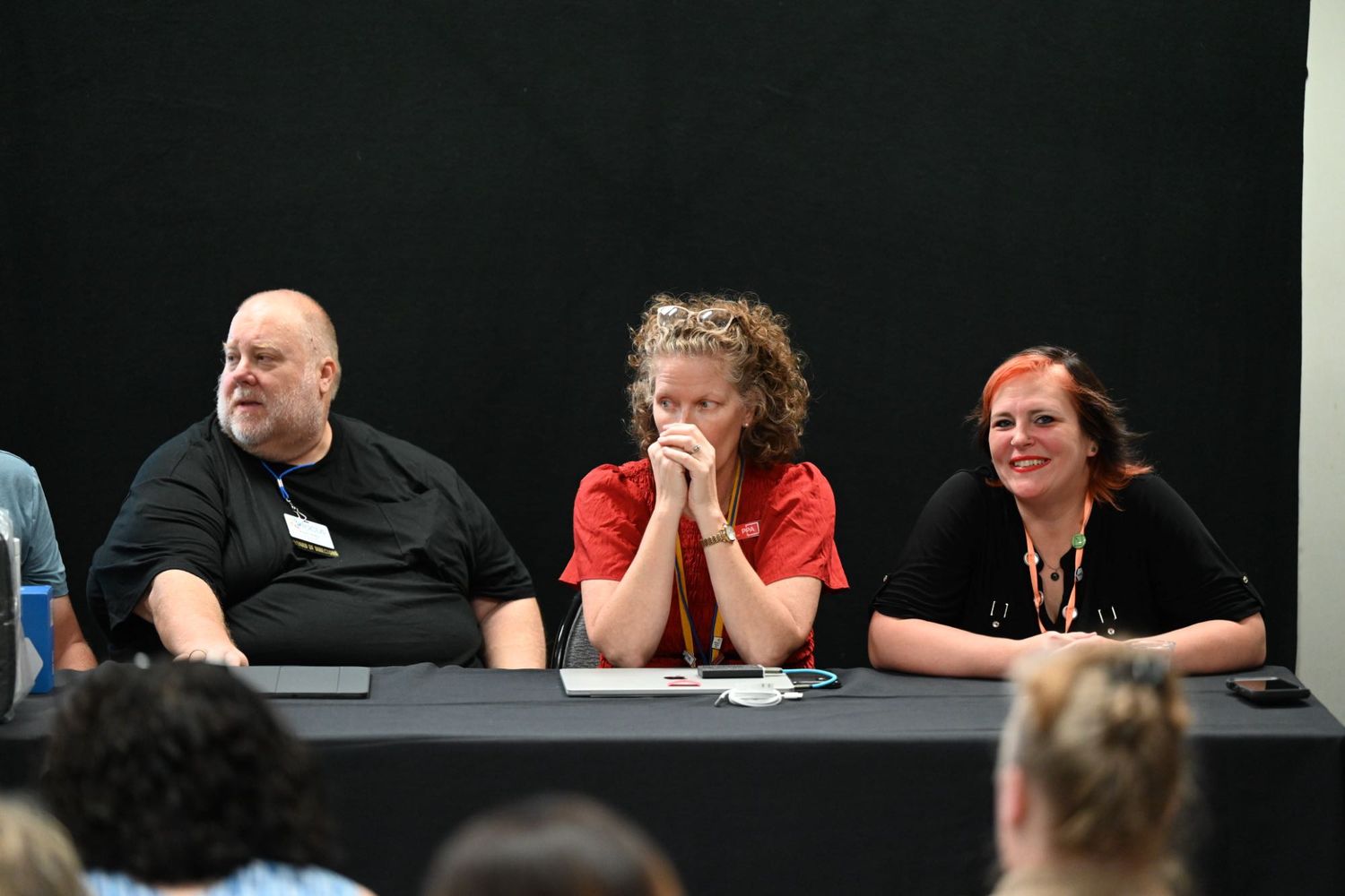 Three adults sit at a panel table during a conference event, with a black backdrop behind them.
