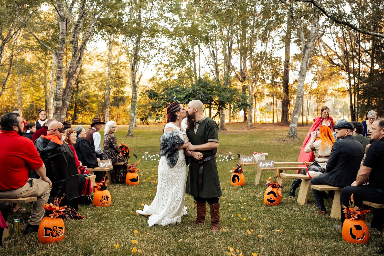 Outdoor autumn Viking wedding ceremony with pumpkin decorations and guests seated on rustic wooden benches.