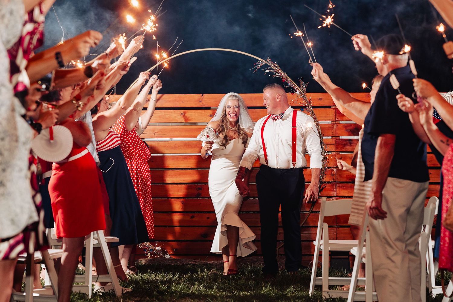 Wedding guests wave sparklers over bride and groom seated in chairs during sparkler send-off at night.
