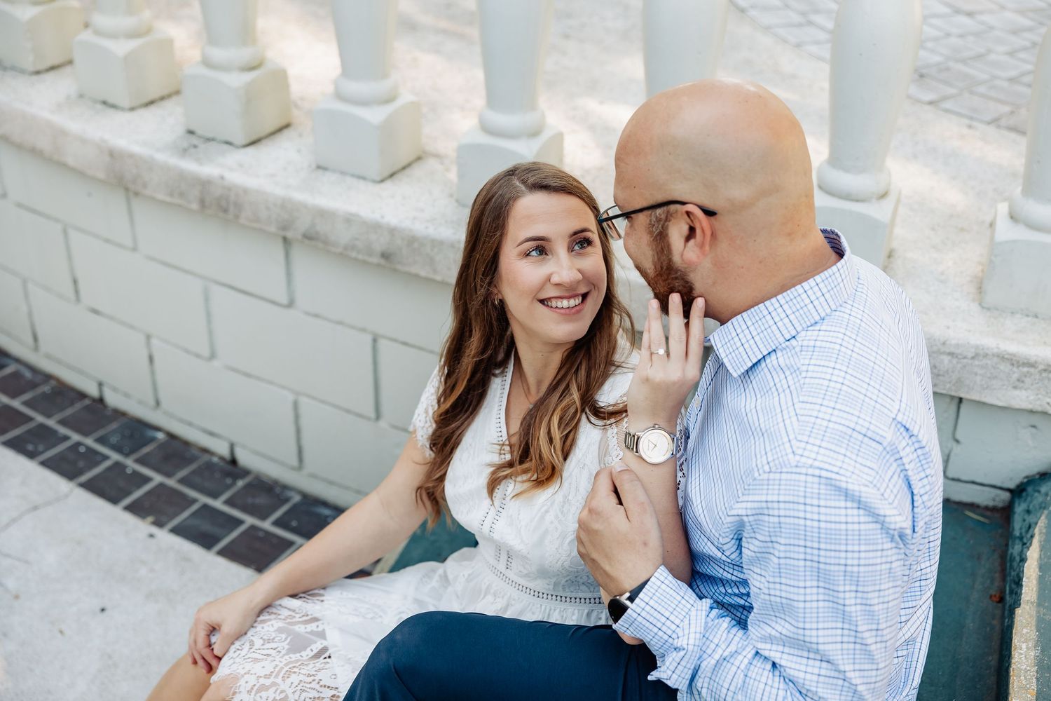 Smiling woman holding mans hand while sitting on steps near white stone columns outdoors.