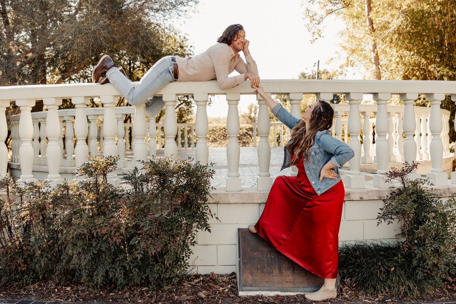A playful couple poses at a white balustrade fence, with the man perched on top holding the woman's hand.