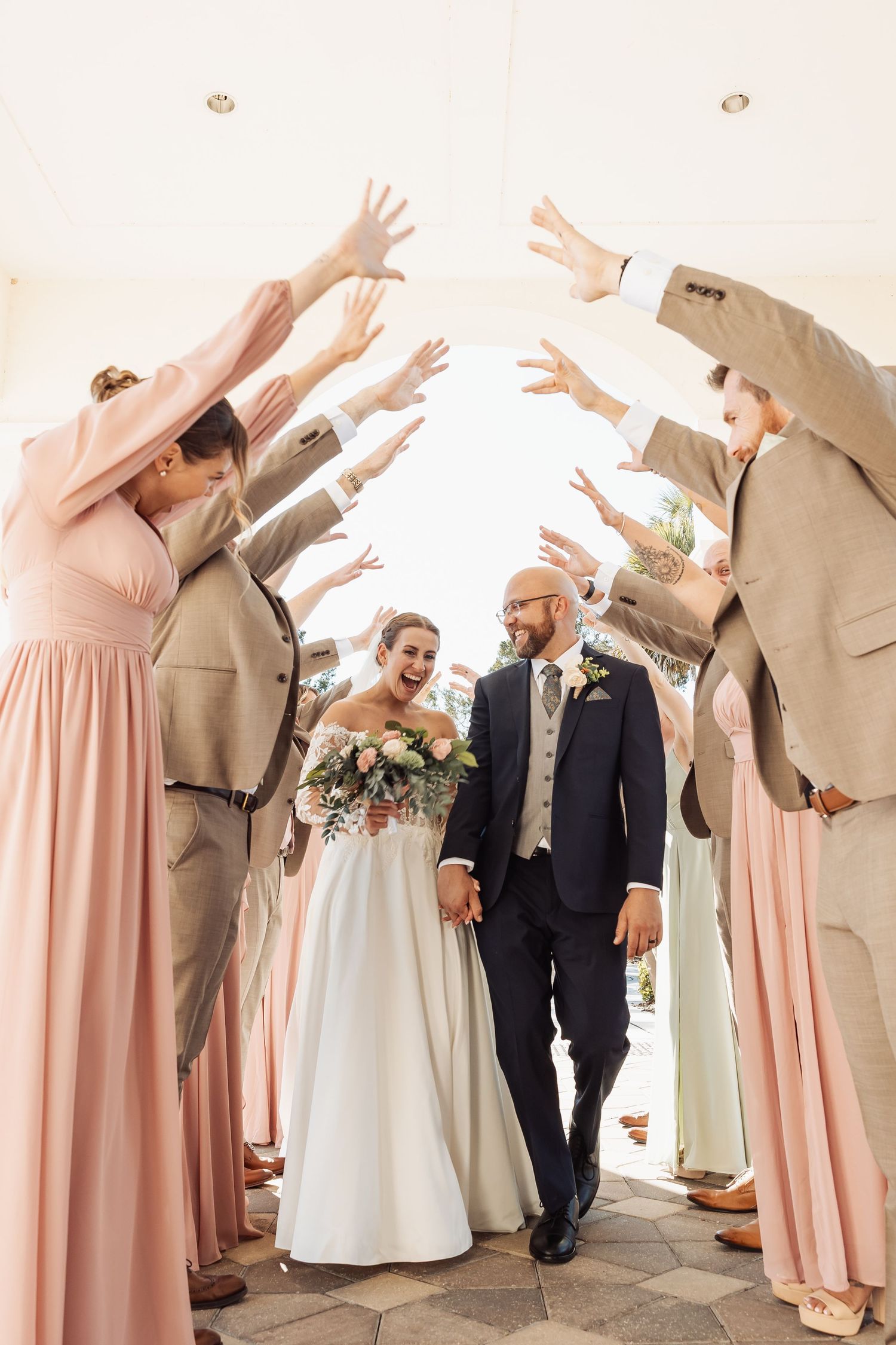Bride and groom walk through arch of raised hands formed by bridesmaids in pink gowns and groomsmen in tan suits.