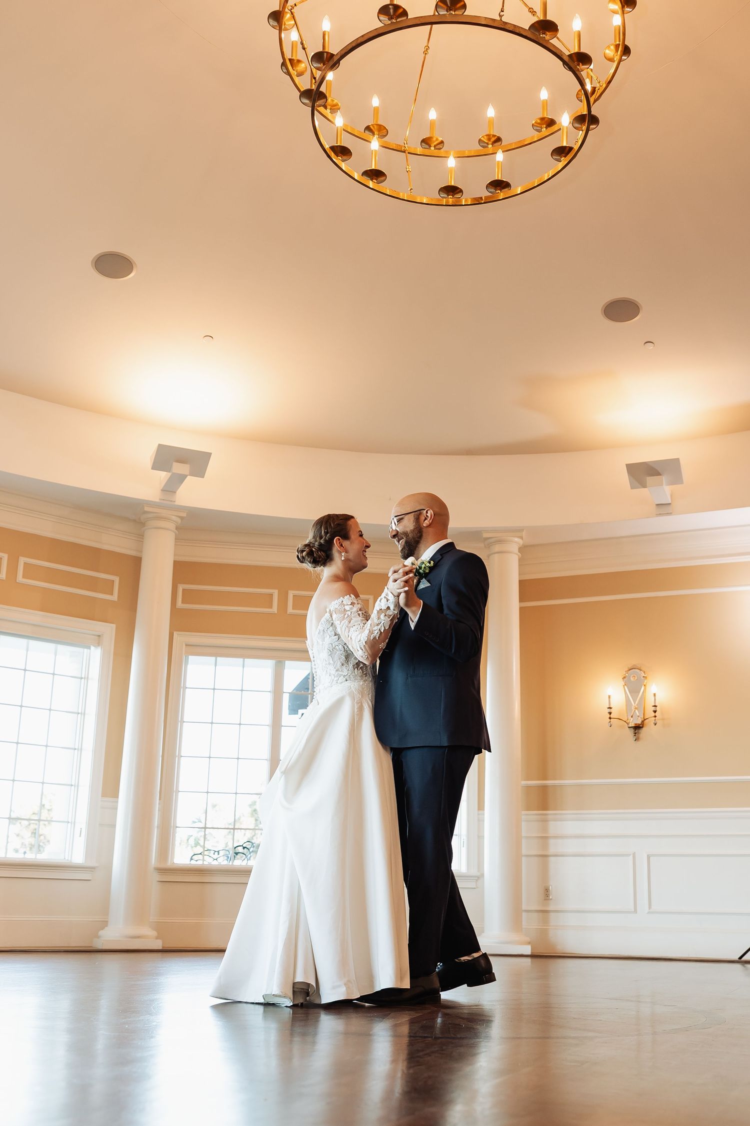 Bride and groom share elegant first dance under ornate chandelier in a warmly lit ballroom.