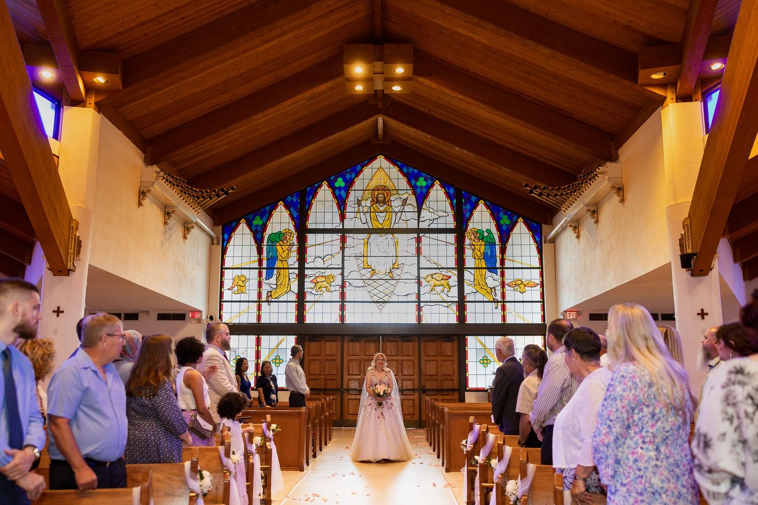 Bride walks down aisle of church with beautiful stained glass windows during wedding ceremony.