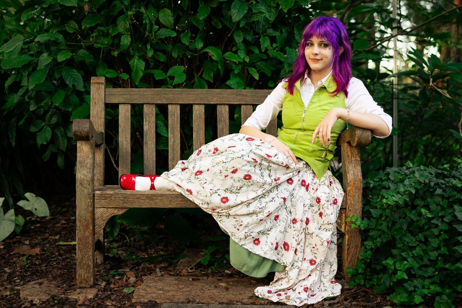 Woman with purple hair in floral skirt and green vest sitting on wooden garden bench surrounded by lush greenery.